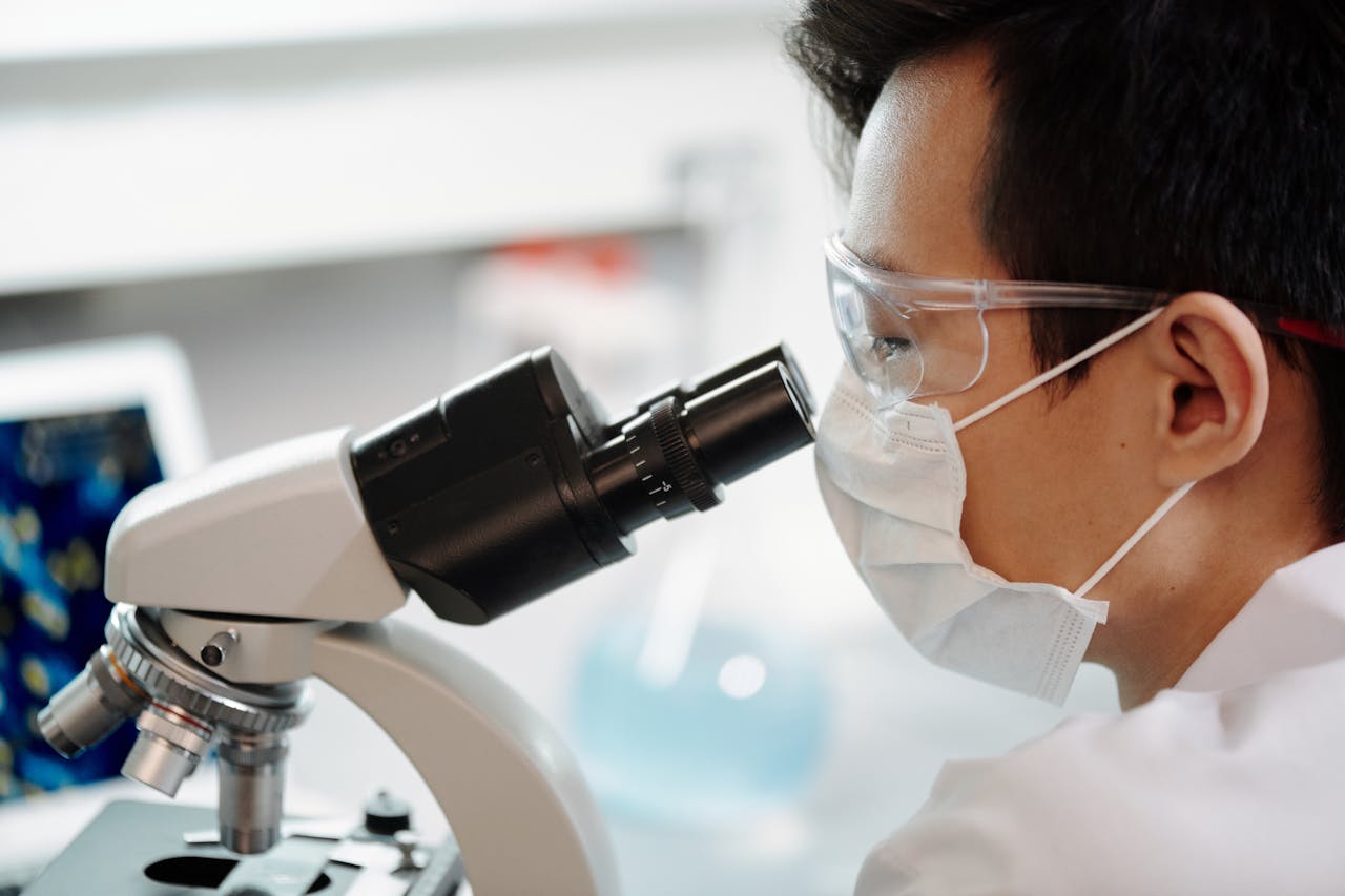 Scientist wearing face mask and goggles examining samples through a microscope in a lab.