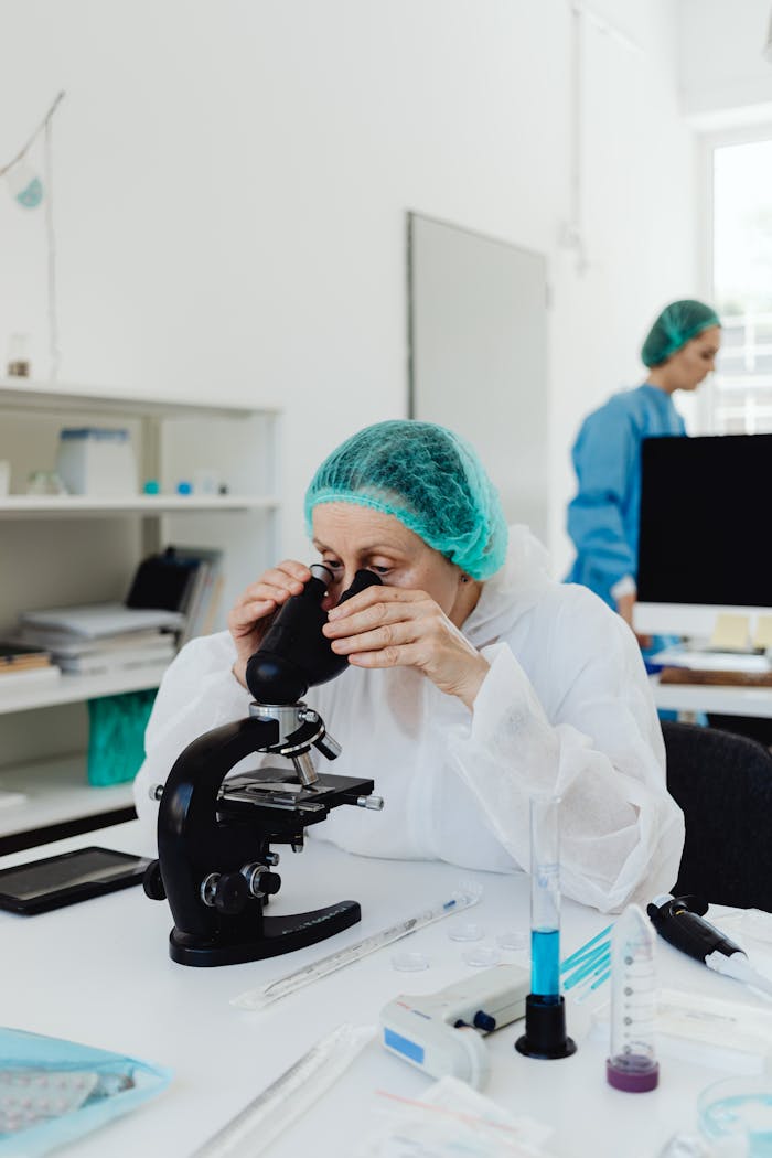 Elderly female scientist in PPE using a microscope in a lab setting.